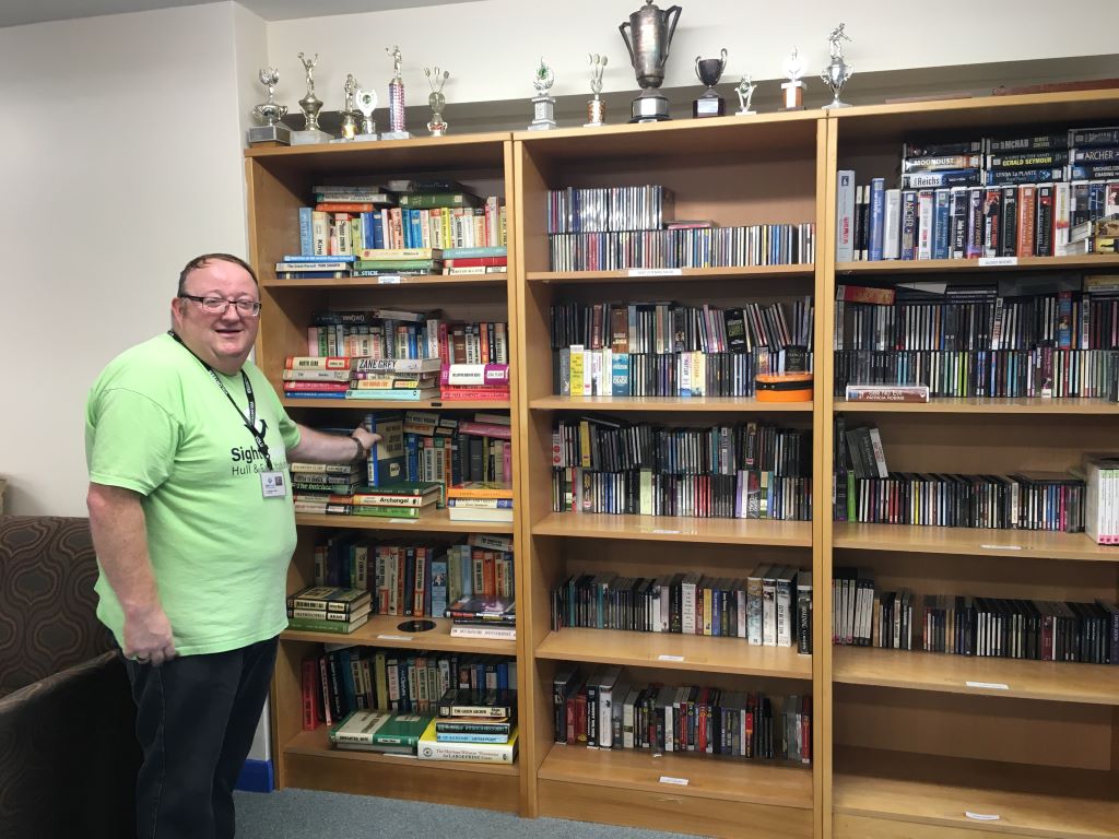 A smiling man stood beside some bookshelves and wearing a green tshirts with the Sight Support logo on it.