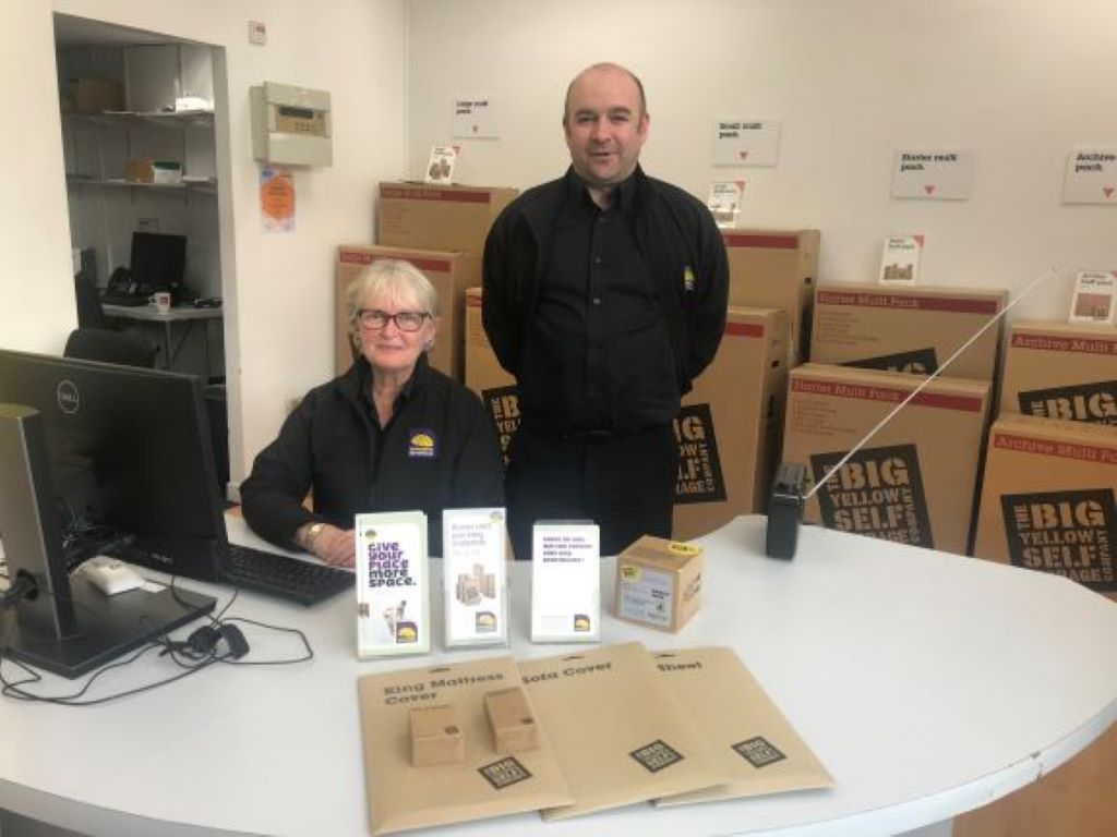 a standing man and seated woman behind a counter with boxes marked Big Yellow Storage behind them.