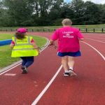 A man and woman running around an athletics track joined at the wrists by a cord.