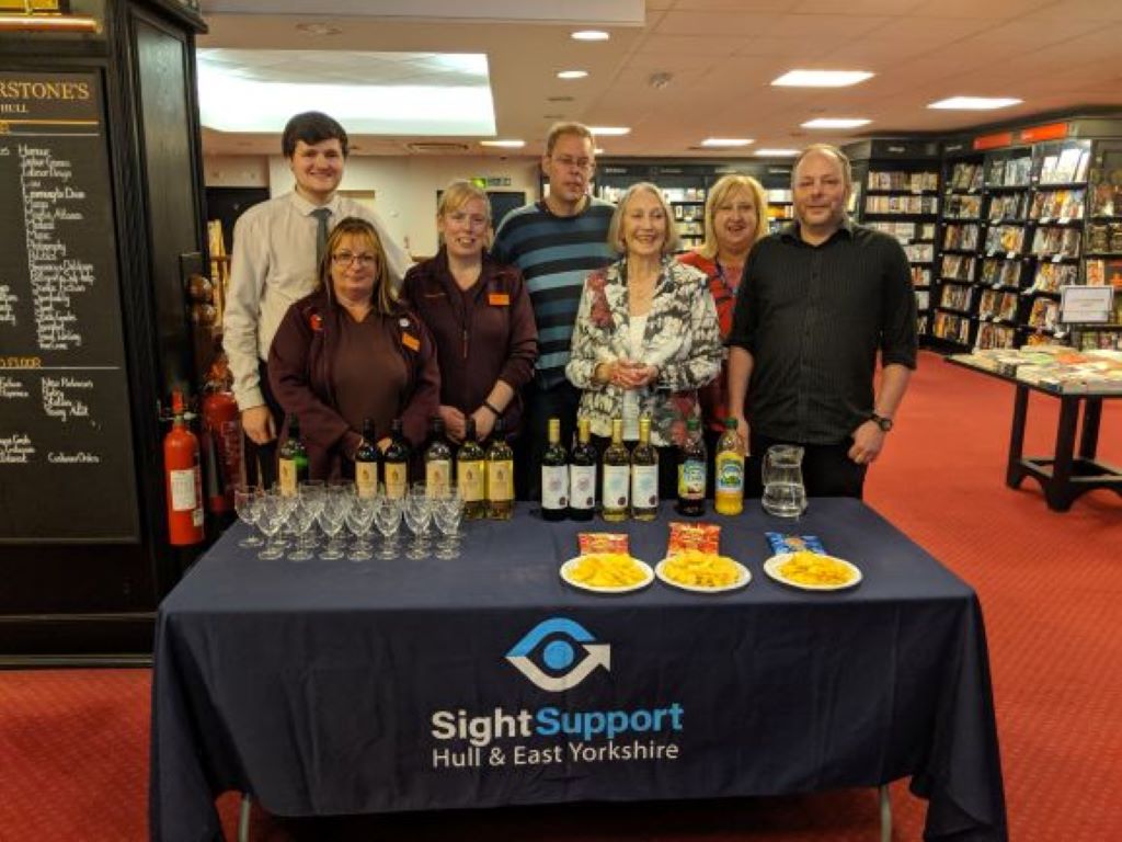 A group of smiling peole standing behidn a table with a tablecloth bearing the Sight Support logo and on top of which are a selcetion of drinks and snacks.