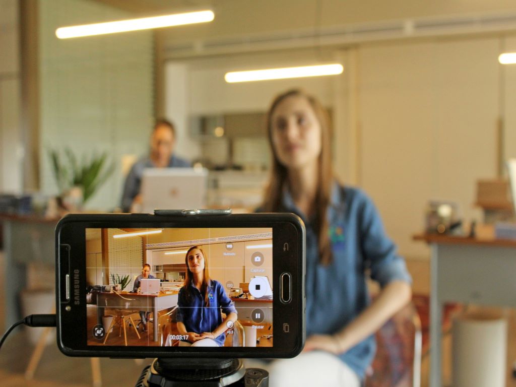 a close up of a mobile phone filming a woman sitting on a stool.