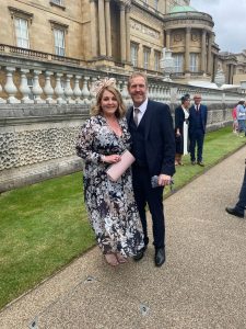 A smiling lady and gentleman standing on the lawns at Buckingham Palace.