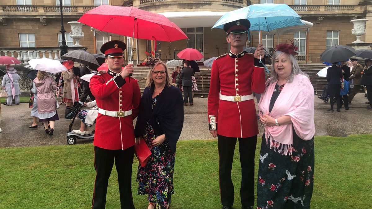 Two smiling ladies standing besides to solders in red and black uniform holding umbrellas.