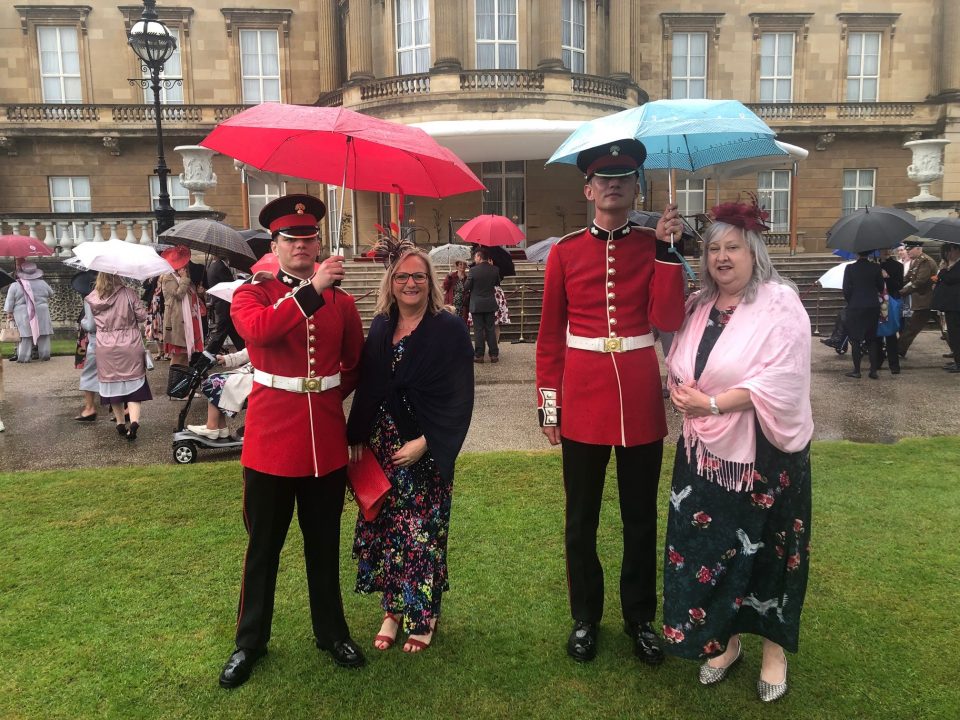 Two smiling ladies standing besides to solders in red and black uniform holding umbrellas.