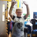 A smiling boy holding up both arms and hands which are covered in paint.