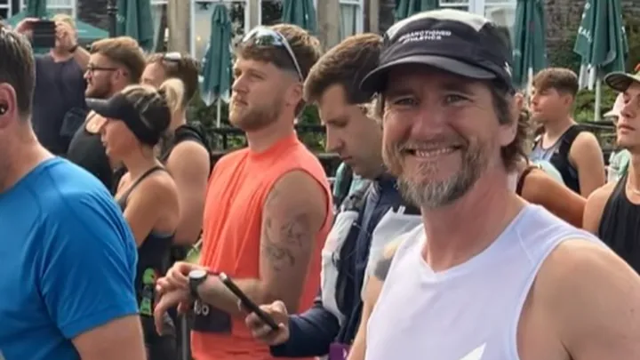 A smiling man wearing a running vest and baseball cap at the start of a running race.