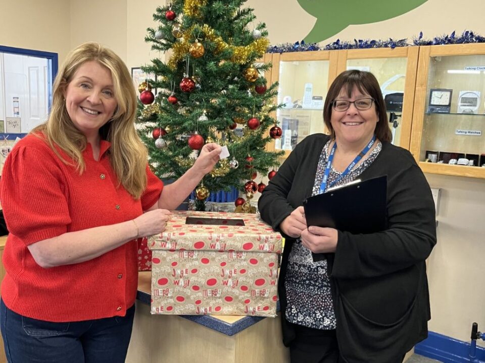 Two smiling ladies standing in front of a Christmas Tree and a large box wrapped in Christmas paper .