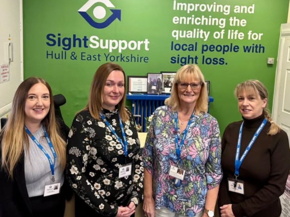 Four smiling ladies standing in front of a wall featuring the Sight Support Hull & East Yorkshire logo